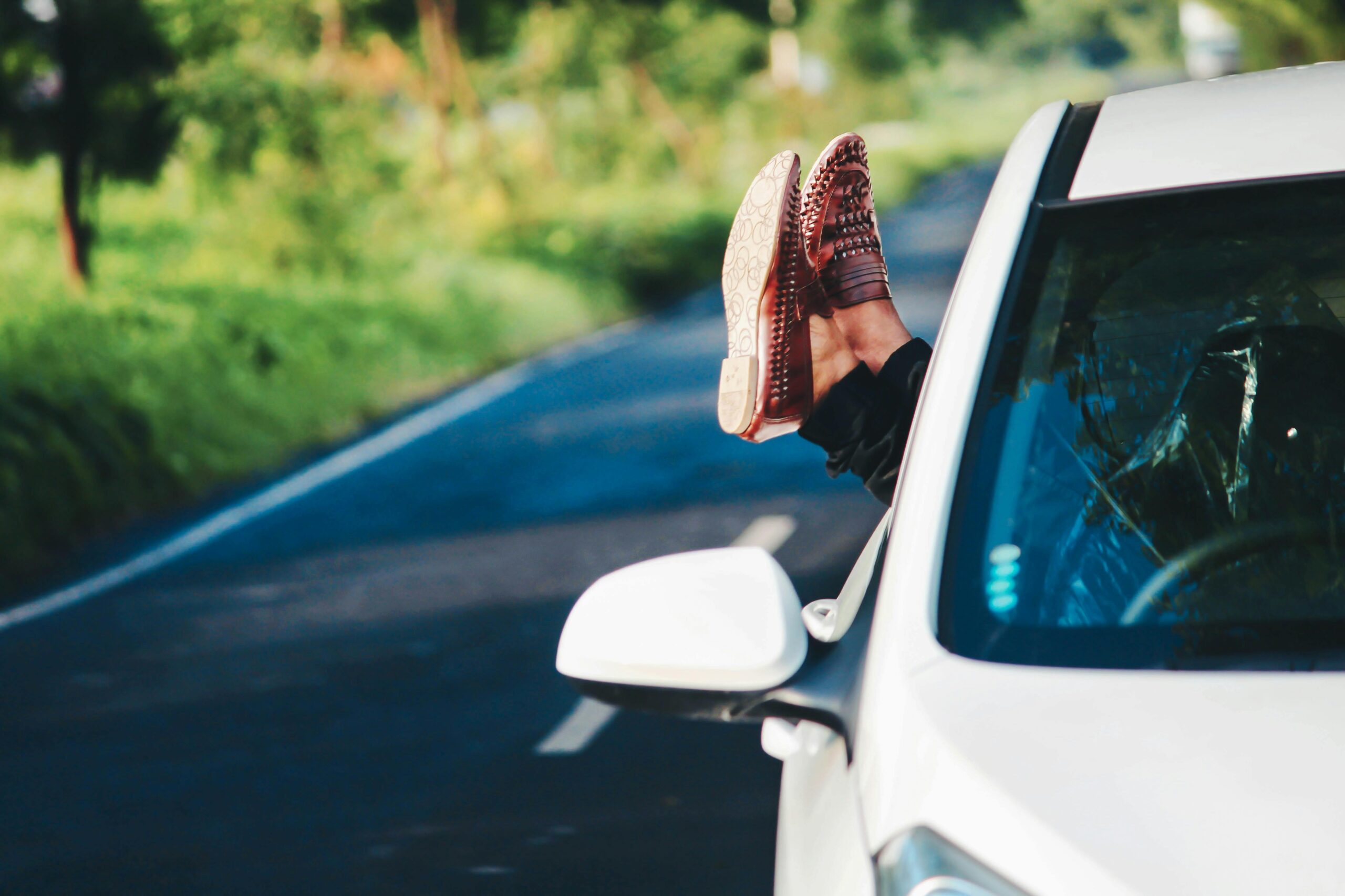 Man relaxing in car, scenic road trip in summer. Feet out window, enjoying countryside.