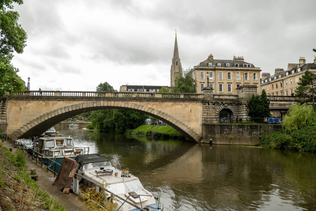 A scenic view of a stone bridge over the river in Bath, England, with moored boats and historic buildings.