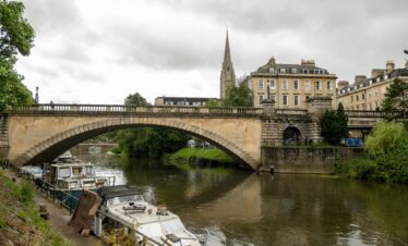 A scenic view of a stone bridge over the river in Bath, England, with moored boats and historic buildings.
