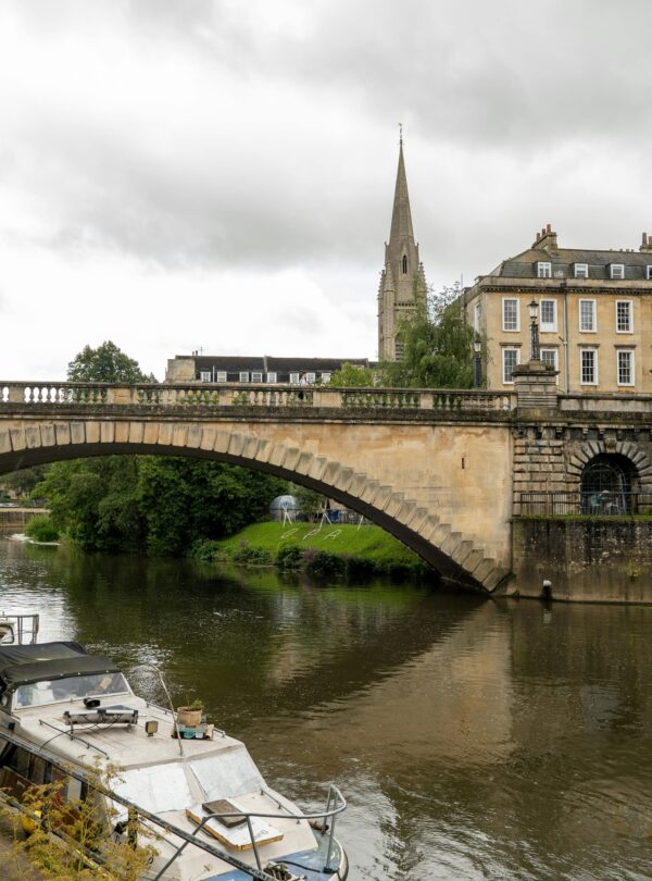 A scenic view of a stone bridge over the river in Bath, England, with moored boats and historic buildings.